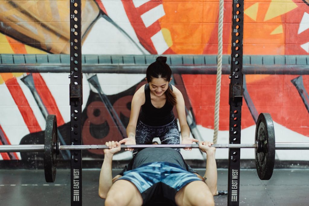 Man lifting weights in gym assisted by woman with colorful mural background.